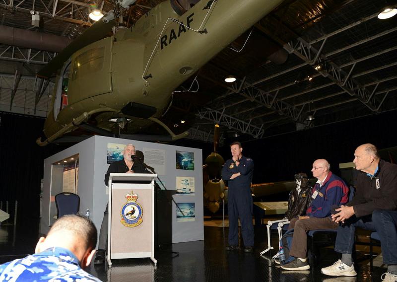 A man speaks indoors at a lectern as people sit facing him, with a military helicopter suspended from the ceiling overhead.