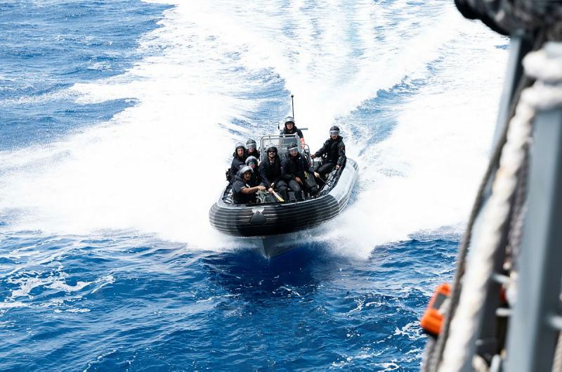 People wearing helmets ride in a small boat across the water, with spray trailing behind.