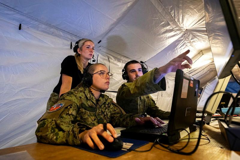 Three Air Force personnel from different countries wear headsets and look at a screen with one person pointing at it.