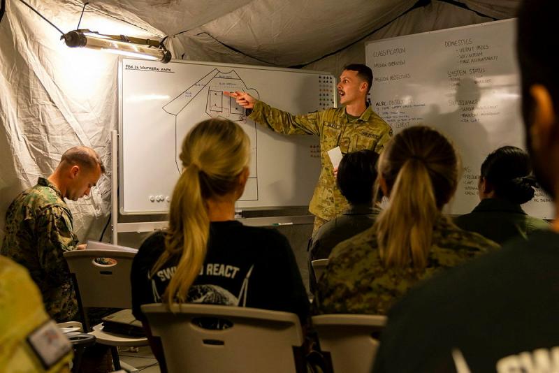 A military member speaks to other personnel about a diagram on a whiteboard.