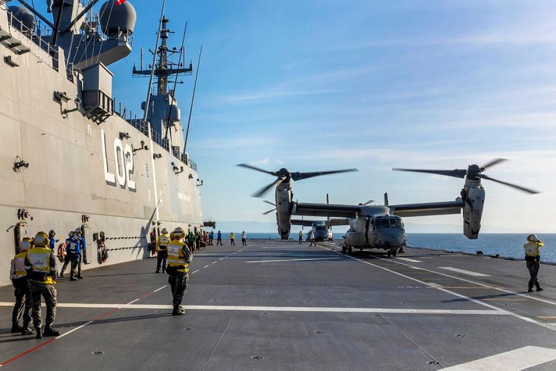 Osprey aircraft sit on the deck of a ship with numerous personnel standing around. 
