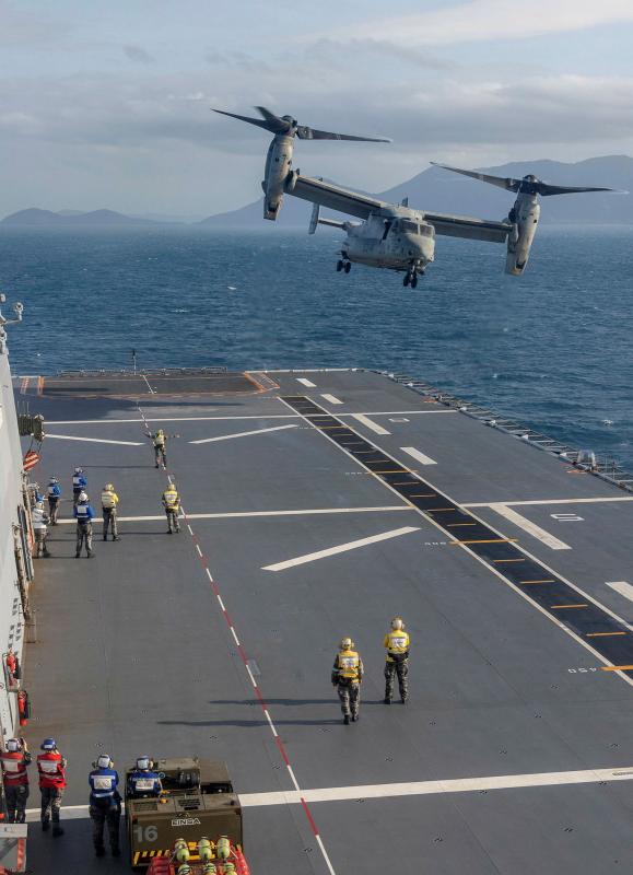 An aircraft hovers above the deck of a ship at sea. People are seen on the deck, looking up.