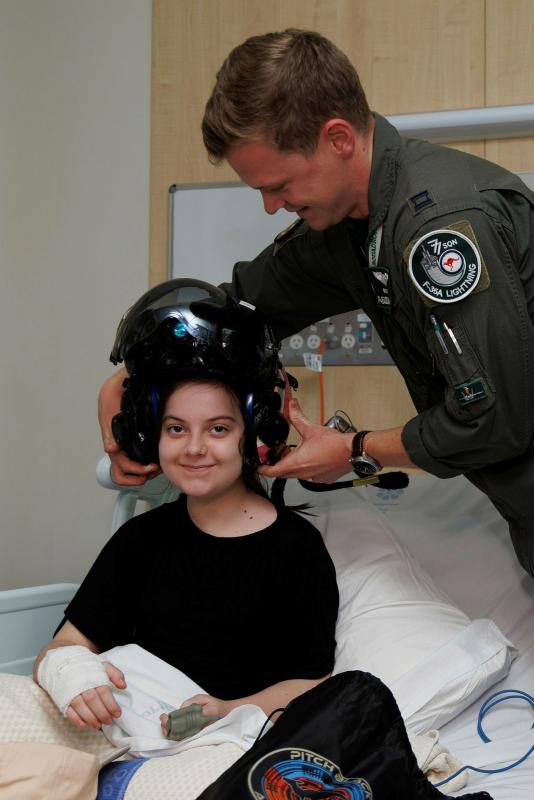 A military man places his pilot helmet on the head of a young girl seated in a hospital bed. 