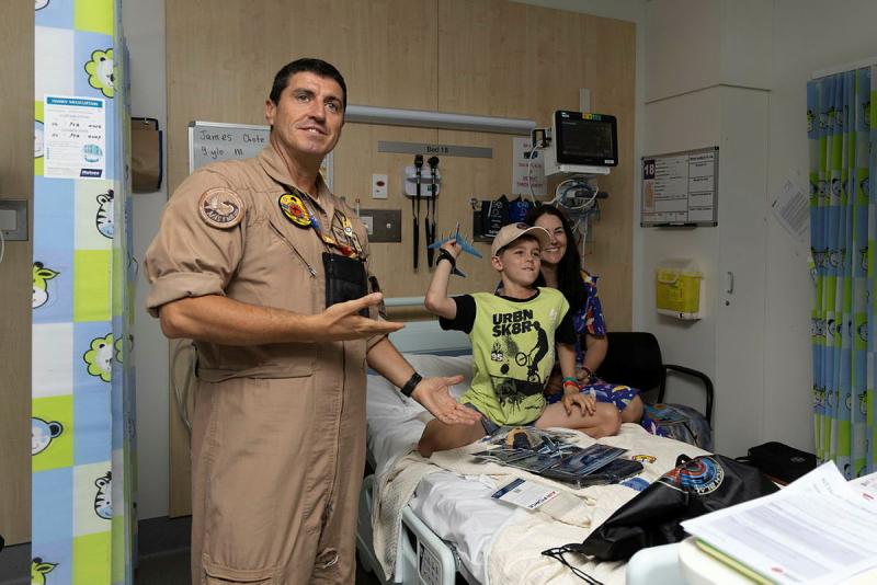 A military man gestures towards a child in a hospital bed about to throw a miniature aircraft. 