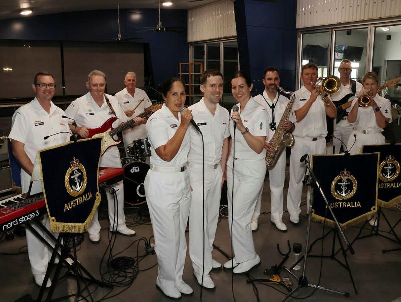Navy personnel holding instruments and microphones perform on a stage.