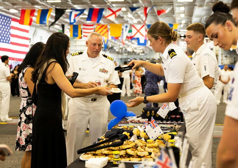 A woman in a white naval uniform uses tongs to place baked goods onto paper plates held by guests.