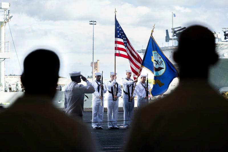 Four people in white naval uniforms stand with flags and rifles on a ship's deck while another salutes nearby.