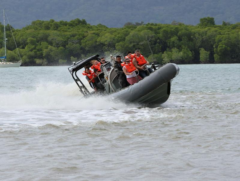 People wearing safety vests join a Navy member on a boat which is spraying water up as it moves through water.