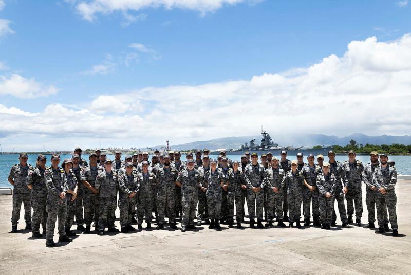 A large group of navy personnel are seen with water and a ship in the background.
