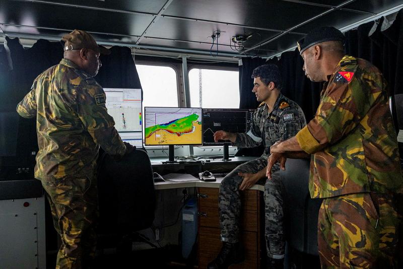 Three people inside a dark room on a ship look at maps on a computer screen.