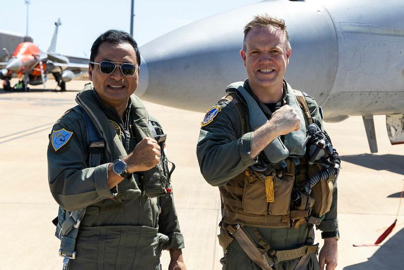 Two military men make the same gesture with their arm in front of an aircraft.