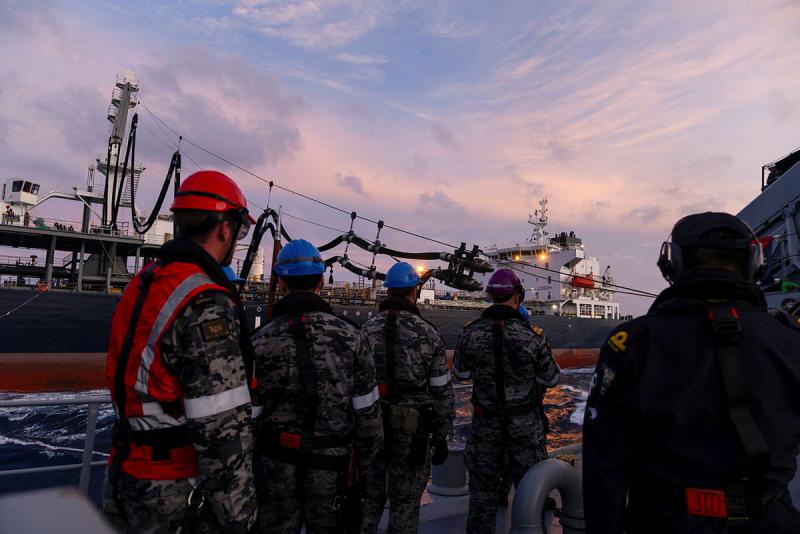 Sailors stand under a pink sky looking out from one ship to another.