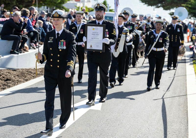 A woman in Navy uniform holds an upraised sword while walking down the street as others follow and spectators watch.