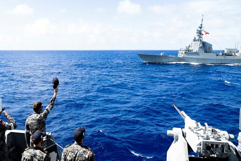 Navy personnel stand on the deck of one ship and wave, across the water, to another.