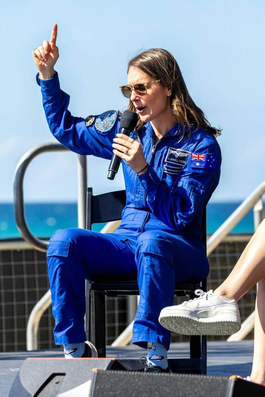 A military member in a blue jumpsuit talks into a microphone and points up at the sky.