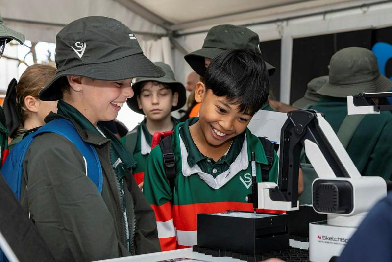 Several young kids inspect a 3D printing machine.