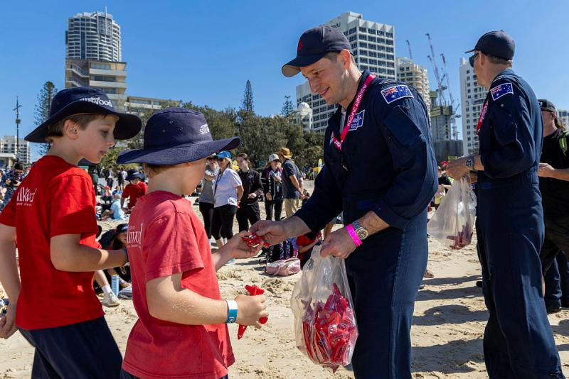 A man hands out gifts to young school students on a beach.