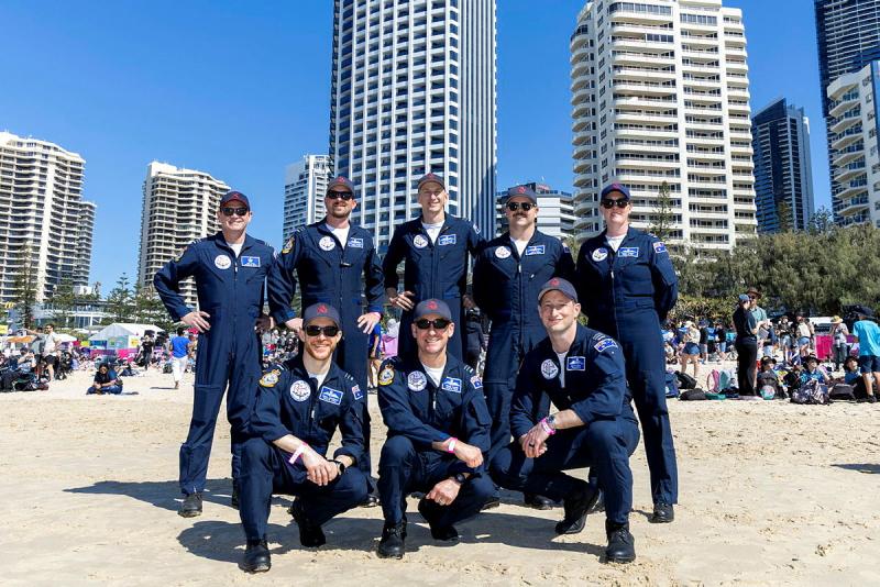 A group of pilots in jumpsuits stand together for a photo on the beach.