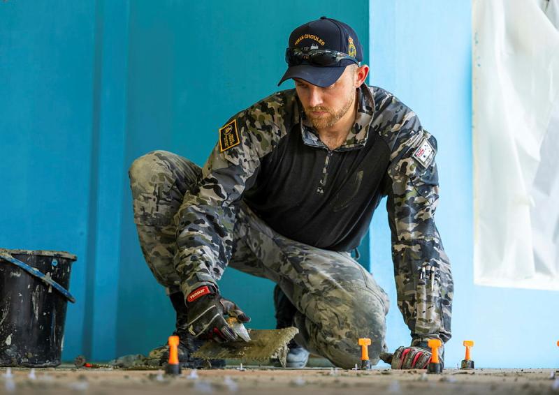 A man in Navy uniform crouches while using a tile trowel.