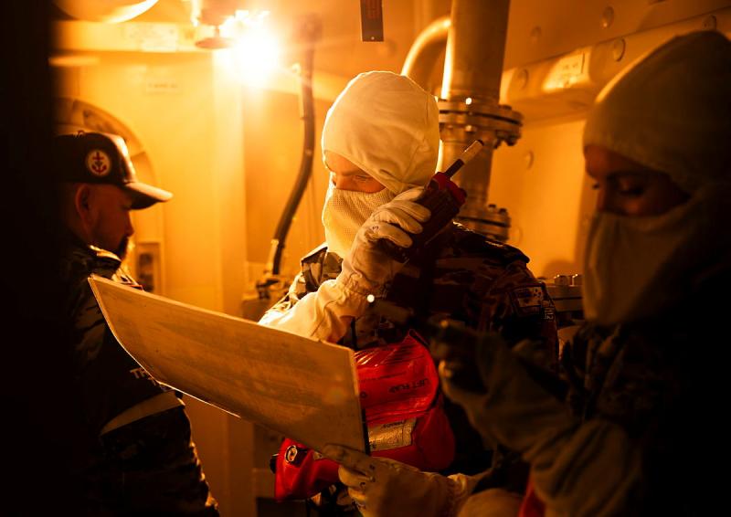 Navy personnel wear protective gear while in a room flooded with orange light. One person is using a walkie-talkie.