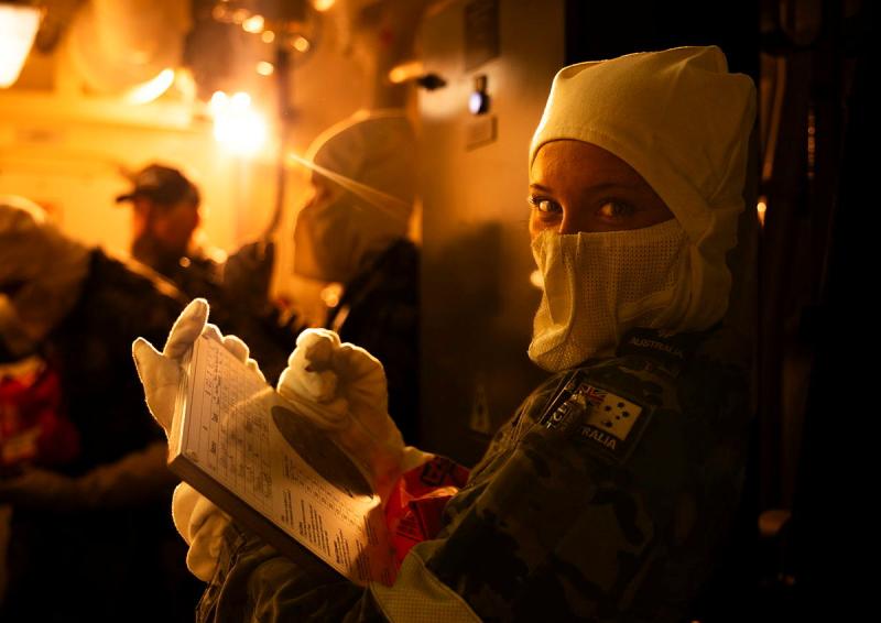A female Navy member is seen holding a document and pen while wearing protective gear in a room with and orange glow.
