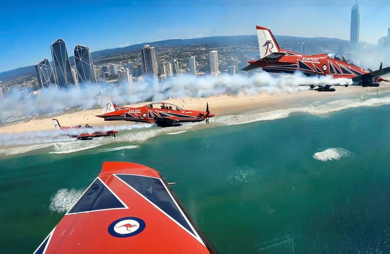 A group of red aircraft fly in formation above a coastal city.