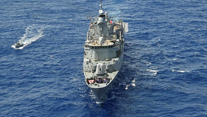 A warship sails on the ocean with people on the foredeck holding Australian, Canadian and New Zealand flags.