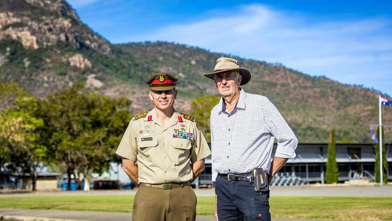 A man in uniform stands next to a man in casual clothes and a broad-brimmed hat, with hills behind them.
