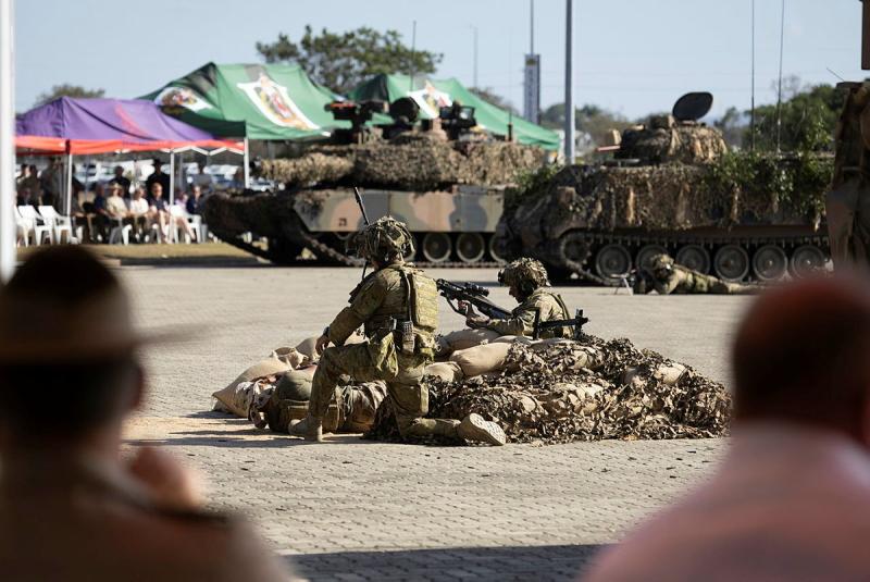 Two soldiers in camouflage sit and kneel with rifles among sandbags, with camouflaged armoured personnel carriers nearby and people watching from under gazebos.