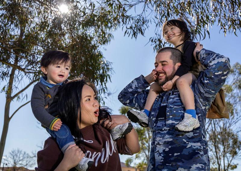 A military man and his wife play with their two children on their shoulders outside.