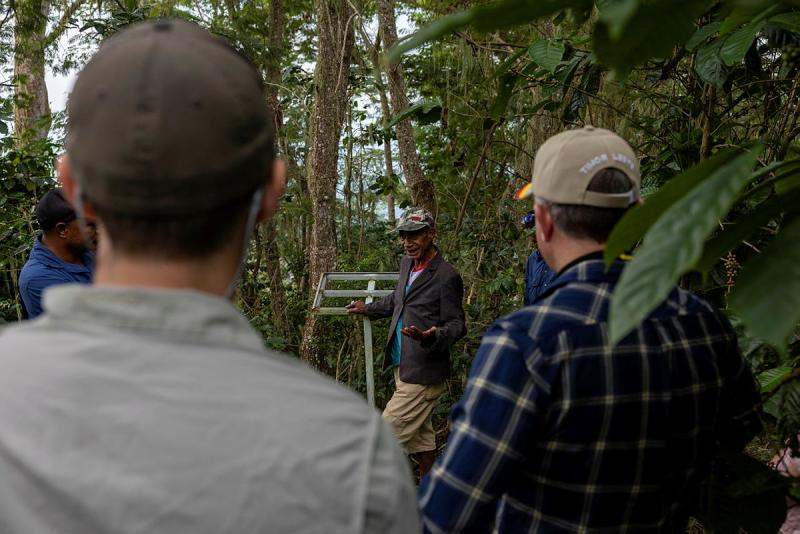 An elderly man in a baseball cap and blazer talks to a group of people in a forested area.