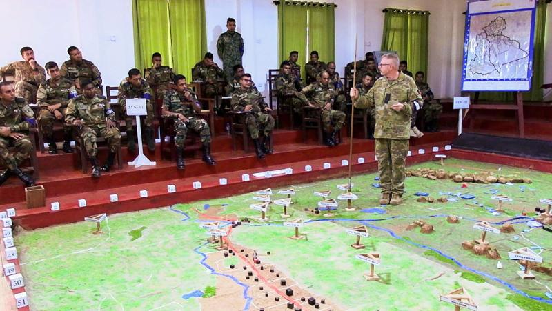 A military man standing on a large map while teaching a small crowd of people.