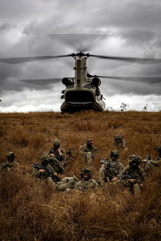Soldiers hide among tall grass, with a helicopter behind them.