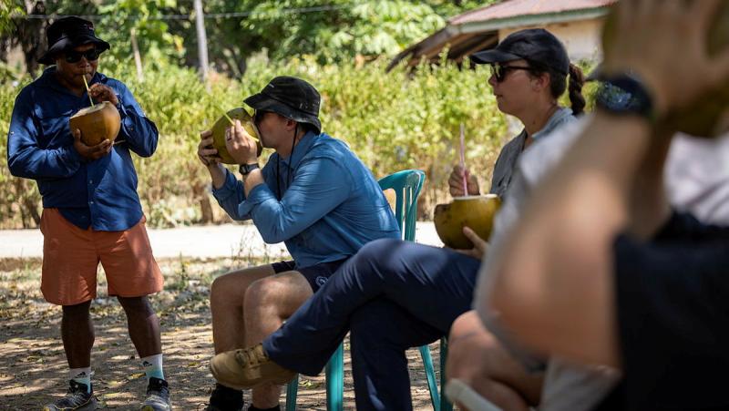 Several people drink water from cut open coconuts while in the shade.
