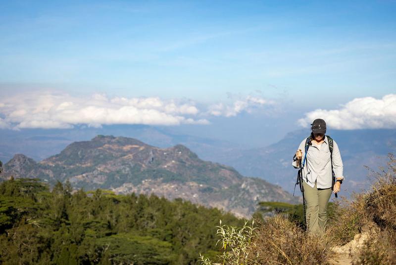 A woman wearing a baseball cap stands on a peak overlooking a valley.