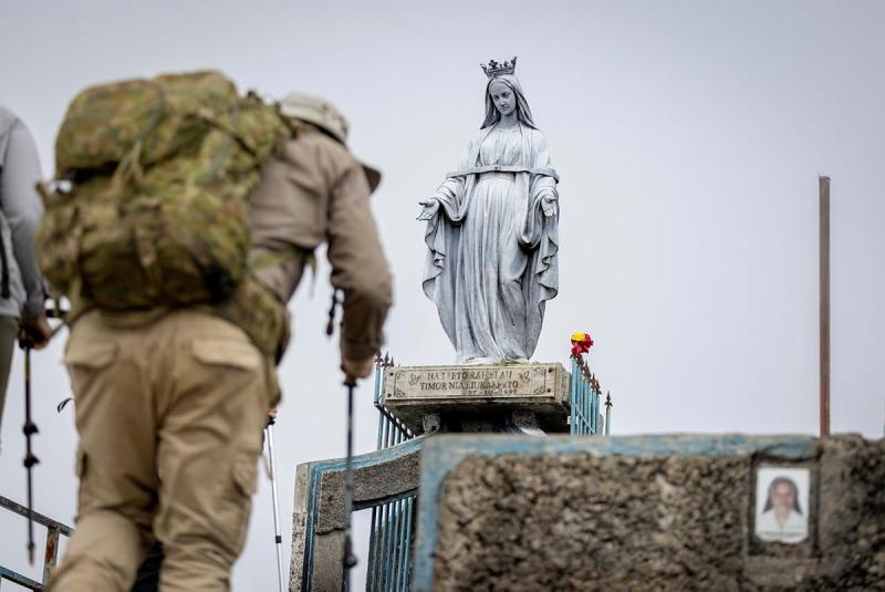 A military member wearing a large pack hikes up to a grey statue of a crowned woman.