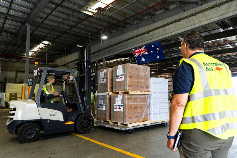 An Australian Aid worker watches a military man lift pallets of Australian Aid in a mini forklift.