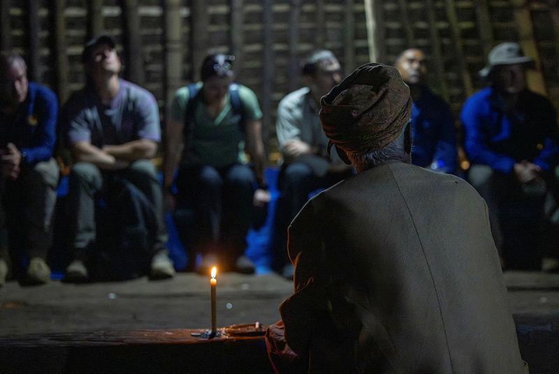 A man in traditional clothing sits opposite a group of people in casual clothing, a candle burning in front of him. 