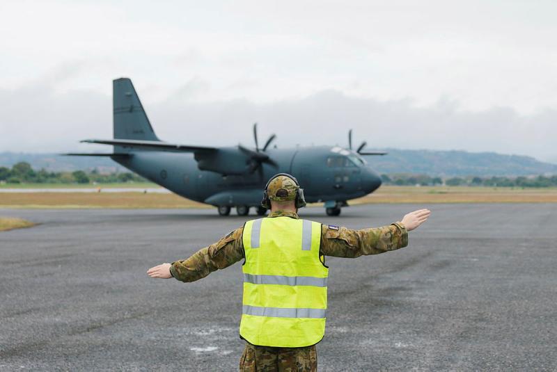 A military member in high-vis signals to a moving aircraft on the tarmac.