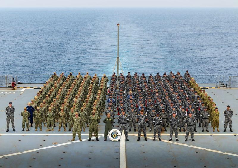 A large group of military people in differing uniforms stand on the upper deck of a ship at sea.