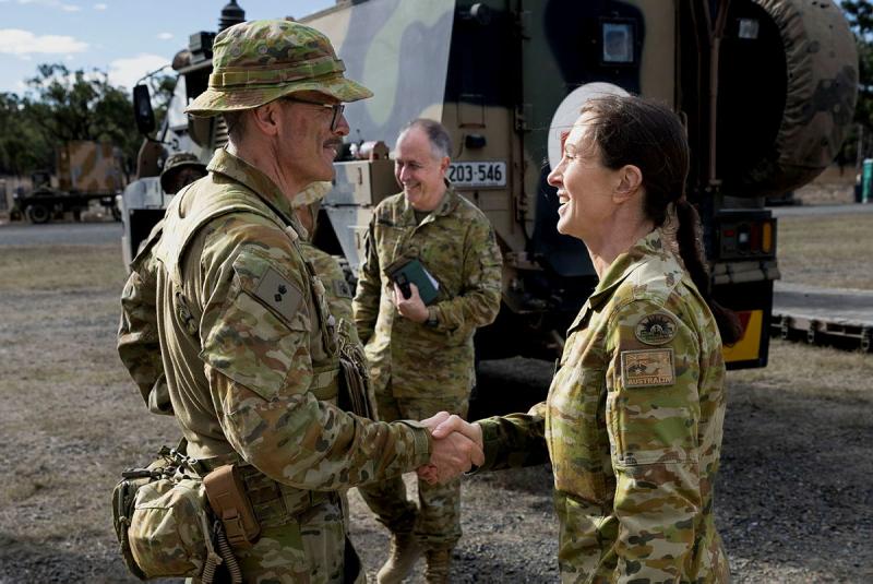 A woman in uniform shakes a man in uniform's hand, with a vehicle in the background.