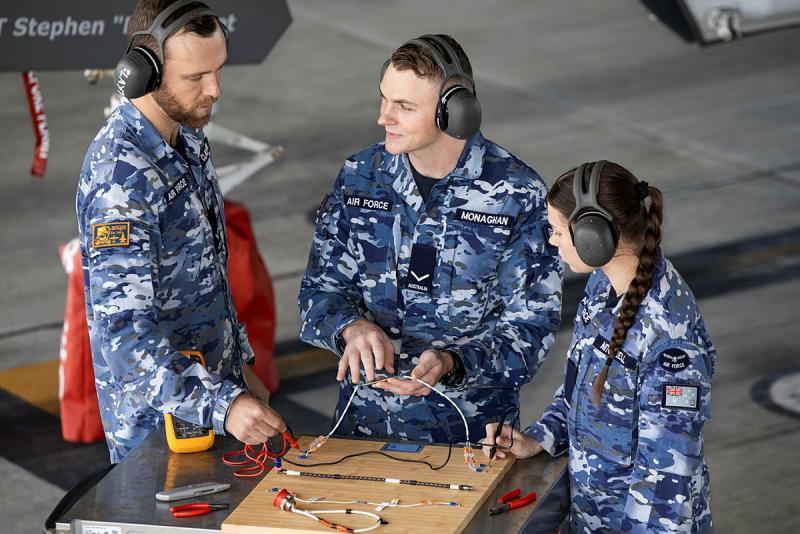 Three military members work on a wooden board affixed with wiring, all while wearing headphones.