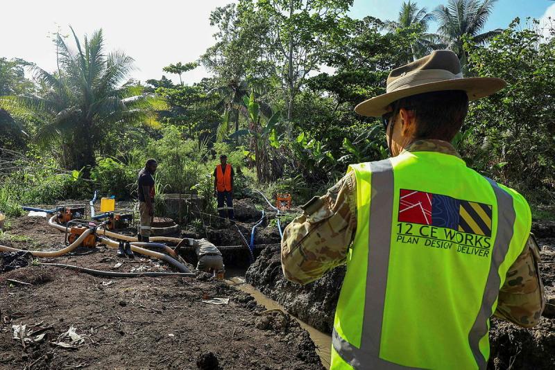 An Army member in a high-vis vest faces PNG locals working on pipes in the ground.