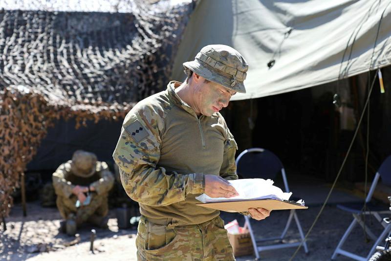 A soldier looks down at his clipboard as he stands in front of a camouflage-covered tent.