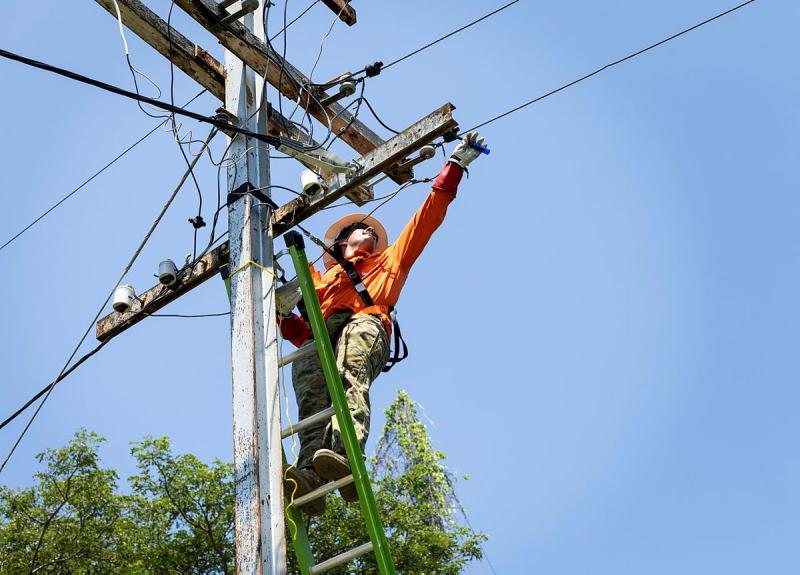 An Army member stands on a ladder which leads against an electricity pole, while reaching out to work on a line.