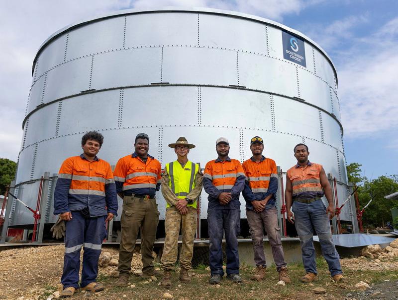 One Army member stands with five PNG workers in front of a large metal tank.