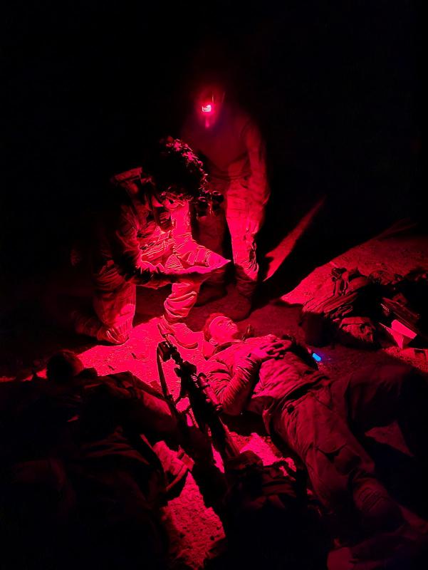 A person in Army uniform crouches down under a red light and looks at another person who is lying on the floor.