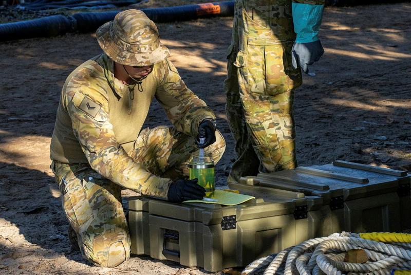 A soldier crouches to look at green liquid in a bottle.