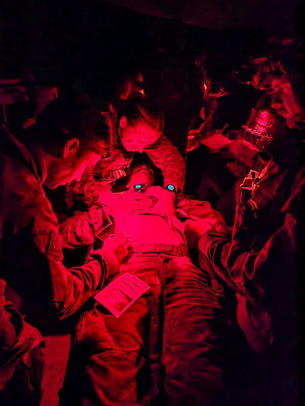 Several military personnel examine a prone military member in the dark, with red lighting.