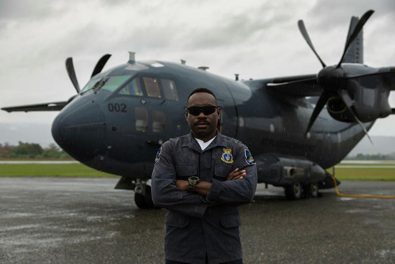 A military man wearing sunglasses stands in front of an aircraft on an overcast day.
